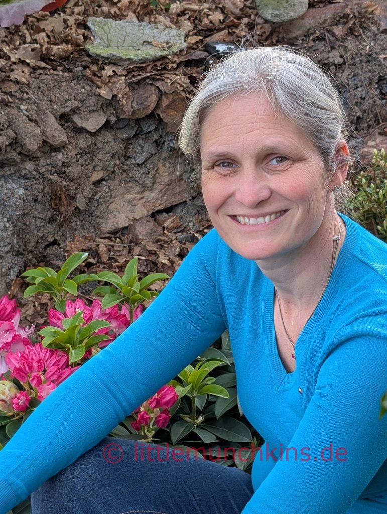 Diana lächelnd im Beet sitzend vor einer rosa Blume und einem Baumstamm