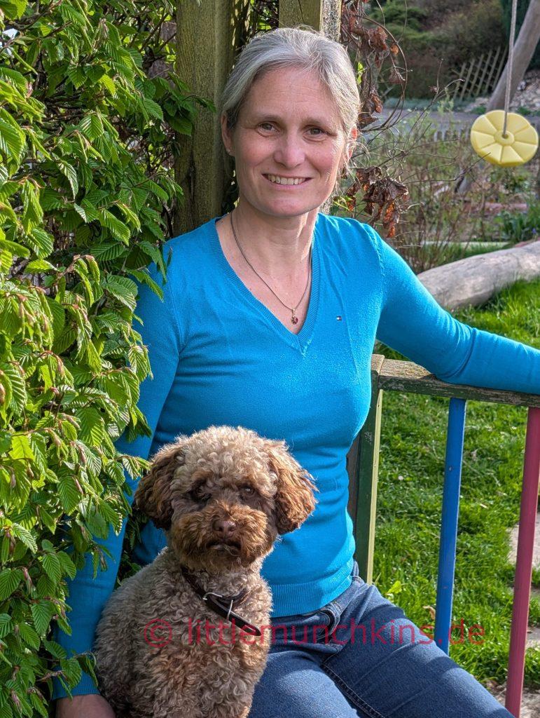 Diana lächelnd sitzt zusammen mit Therapie Hund Holly auf einem Stuhl vor einem buntem Zaun